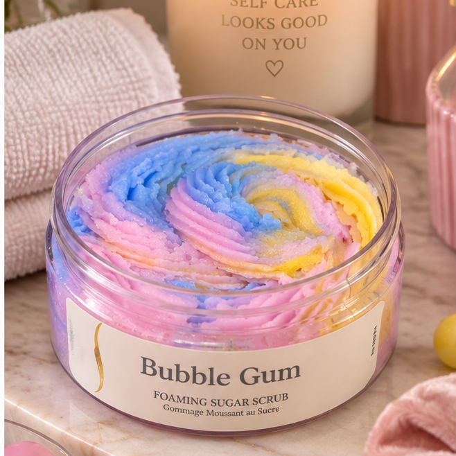 A clear round jar of Bubble Gum foaming sugar scrub sits on a marble surface, filled with pastel pink, blue, and yellow swirled scrub. The jar has a cream label with gold text.