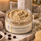 A jar of Caramel Espresso Foaming Sugar Scrub sits on a marble tray in a styled bathroom setting with a lit candle, soap dispenser, dried flowers, coffee beans, and a wooden brush.
