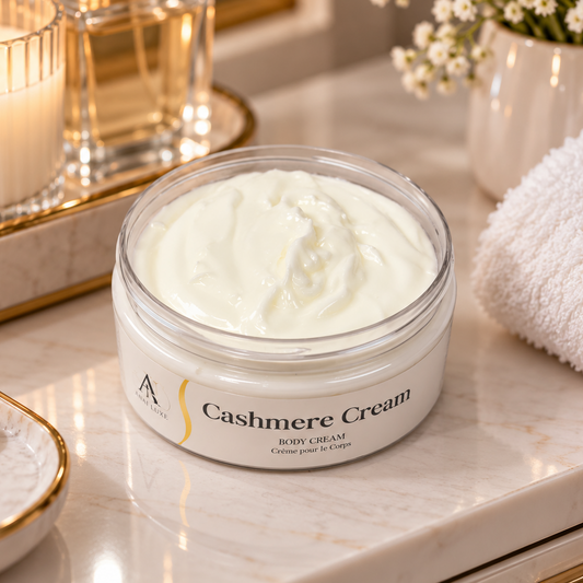 A clear jar of A. Luxe Cashmere Body Cream filled with thick white cream, set on a marble vanity tray with a lit candle, gold-rimmed glass bottles, a white towel, and small white flowers in the background.