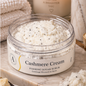 A clear round jar of Cashmere Cream Foaming Sugar Scrub sits on a marble tray, filled with white sugar scrub speckled with black seeds. The scene is styled with a lit candle, folded towels, and butter in the background.