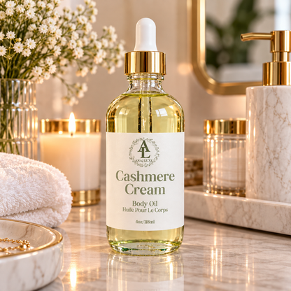 A bottle of Annuluxe Cashmere Cream body oil with a white dropper cap and gold collar, set on a marble bathroom counter with candles, flowers, and soap dispensers in the background.