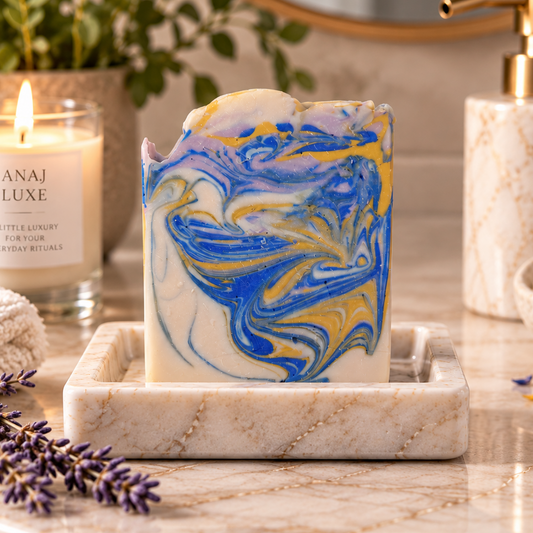 A marbled handmade soap bar with swirls of blue, yellow, and white sits in a marble soap dish on a bathroom counter. A lit candle labeled 'ANAJ LUXE' is to the left, with a gold pump bottle, plants, and lavender sprigs in the background and foreground.