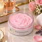 A jar of pink body cream labeled 'Enchantment' sits on a marble surface surrounded by spa props including a candle, perfume bottle, flowers, lavender, and a folded towel.