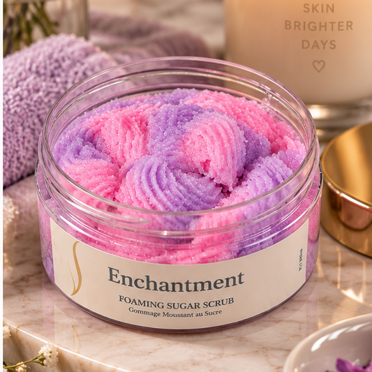 A jar of Enchantment Foaming Sugar Scrub filled with pink and purple whipped scrub, styled on a marble surface with a lit candle, dried flowers, and a purple towel in a spa-like setting.
