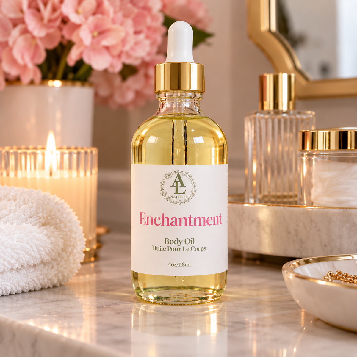 A bottle of Enchantment Body Oil by Lavender Lane stands on a marble countertop in a spa-like bathroom setting, surrounded by a lit candle, pink flowers, a towel, and other gold-and-clear toiletry items.