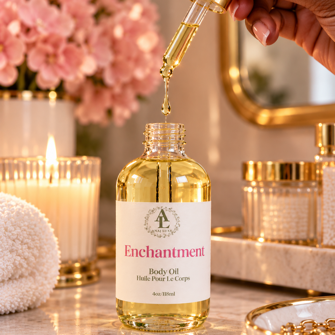 A hand holds a gold dropper above a clear bottle of Enchantment Body Oil, with golden oil dripping into the bottle. The bottle has a white label with pink text and a gold crest logo.
