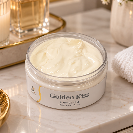 An open jar of Golden Kiss Body Cream sits on a marble vanity surface, surrounded by a lit candle, gold-toned bottles, a folded white towel, and small flowers in the background.