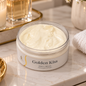 An open jar of Golden Kiss Body Cream sits on a marble vanity surface, surrounded by a lit candle, gold-toned bottles, a folded white towel, and small flowers in the background.