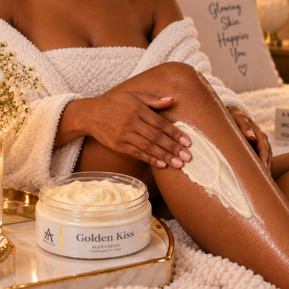 A woman in a white robe applies a thick cream body butter from a jar labeled 'Golden Kiss' to her leg in a cozy bedroom setting with candles and pillows.