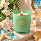 A mint green glass candle jar with a gold monogram logo, lit and styled on a marble tray with seashells, blue crystals, tropical leaves, and a perfume bottle in the background.