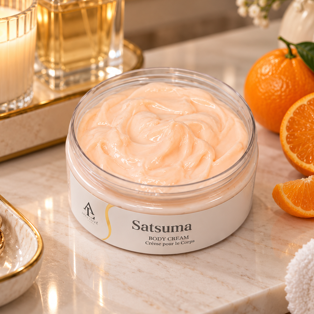 A jar of Satuma Body Cream sits on a marble countertop in a styled bathroom setting, surrounded by oranges, a lit candle, and a white towel.