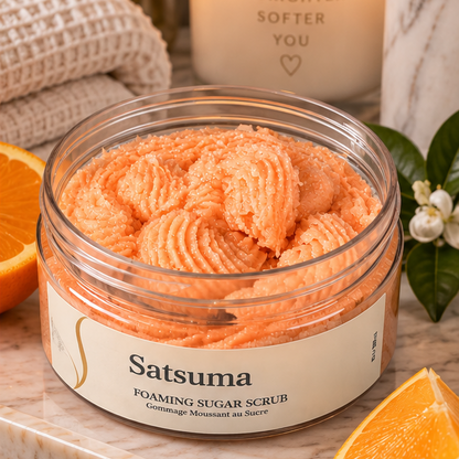 A clear jar of Satuma Foaming Sugar Scrub filled with peach-colored scrub, styled on a marble surface with orange slices, white flowers, a folded knit towel, and a lit candle in the background.