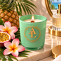 A mint green glass candle jar with a gold monogram logo and wreath design, lit with a small flame. The candle is styled on a marble tray with tropical flowers, a pineapple, a halved passion fruit, and a gold bottle in the background.