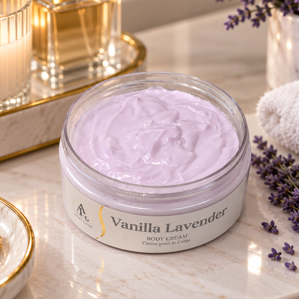 A jar of lavender body cream sits on a marble countertop with spa-style props, including a lit candle, gold-rimmed dishes, a towel, and lavender sprigs. The jar is clear with pale lavender cream visible inside and a label reading 'Vanilla Lavender'.