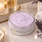 A jar of lavender body cream sits on a marble countertop with spa-style props, including a lit candle, gold-rimmed dishes, a towel, and lavender sprigs. The jar is clear with pale lavender cream visible inside and a label reading 'Vanilla Lavender'.