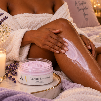 A woman in a white robe applies a light lavender body cream from a jar labeled 'Vanilla Lavender' while sitting on a cozy bed. The scene is warmly lit with candles, lavender flowers, and soft bedding.