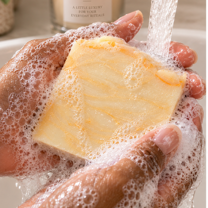 A hand holds a rectangular bar of pale yellow soap under running water from a brass faucet, with lathered foam around it. A candle labeled ANAJ LUXE sits blurred in the background near a plant.