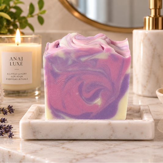 A handcrafted rectangular soap bar with a pink, purple, and white marbled swirl sits on a white marble tray on a bathroom counter. In the background, a lit candle labeled 'ANAJ LUXE' is visible alongside a gold-finished soap dispenser, a mirror, and a rolled towel.