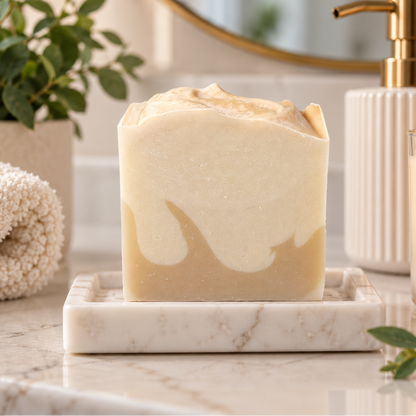 A rectangular bar of handmade soap with a cream and light tan marbled swirl sits on a white marble tray on a bathroom counter. A lit candle labeled 'ANAJ LUXE' and a white ribbed soap dispenser are visible in the background, along with a folded towel and green leaves.