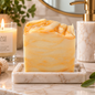 A marbled yellow and cream soap bar sits on a marble tray on a bathroom counter, styled with a lit candle, soap dispenser, brush, and greenery. The candle label reads 'ANAJ LUXE'.